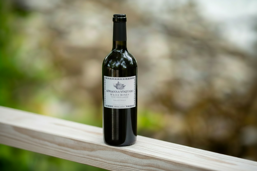 A bottle of Adrianna Vineyard White Bones chardonnay sitting on a wooden surface with trees and ocean in the background.