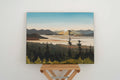 A Fiona Dalrymple landscape painting of the Pacific Northwest with a mountain lake at sunset, displayed on a rustic wooden shelf against a white wall.
