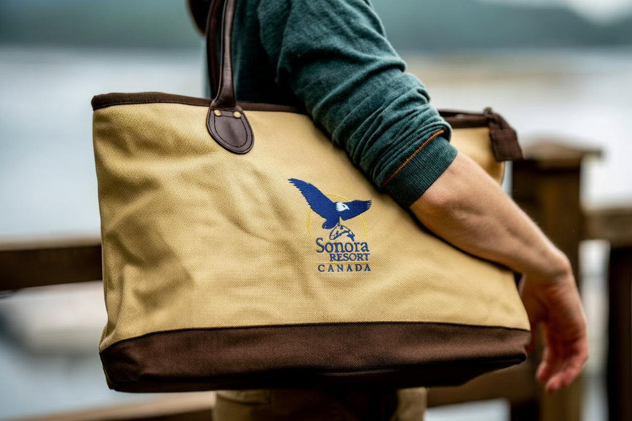 Beige tote bag with a Sonora Resort logo, held by a person in front of a body of water.