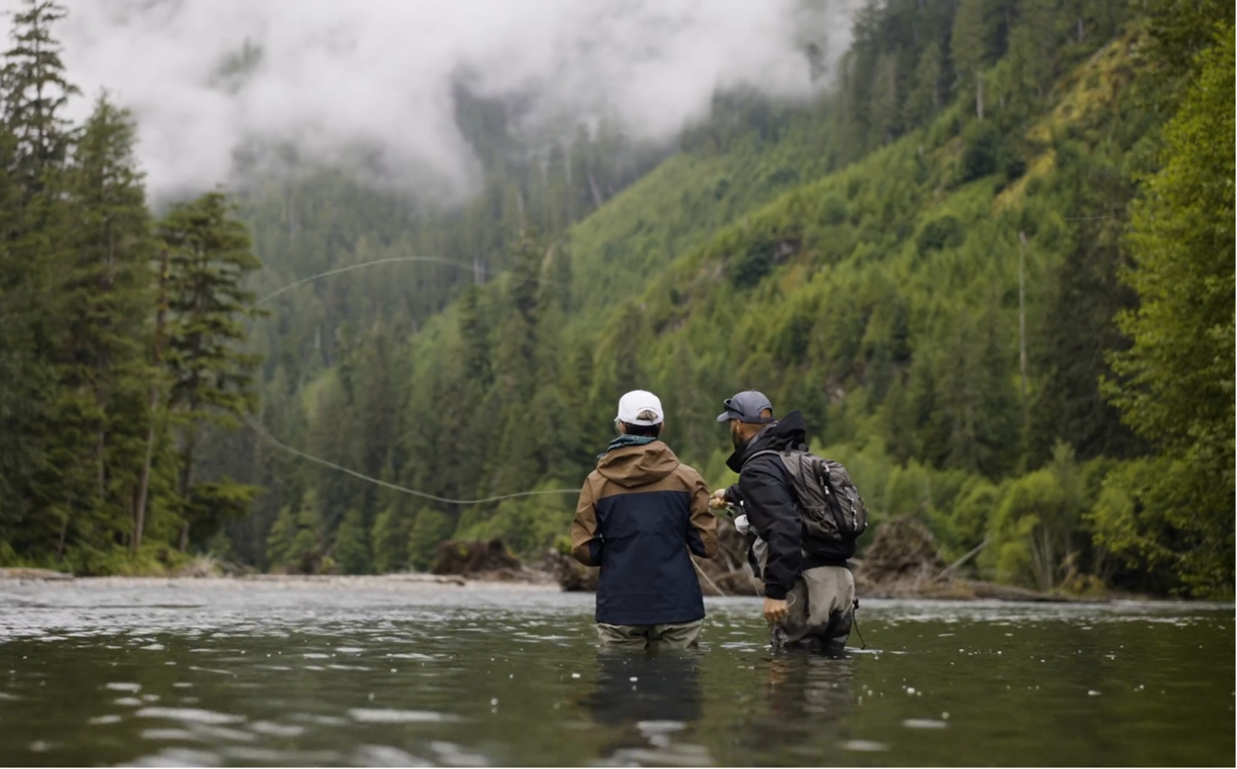 Two people fly fishing in a forested area with misty mountains.