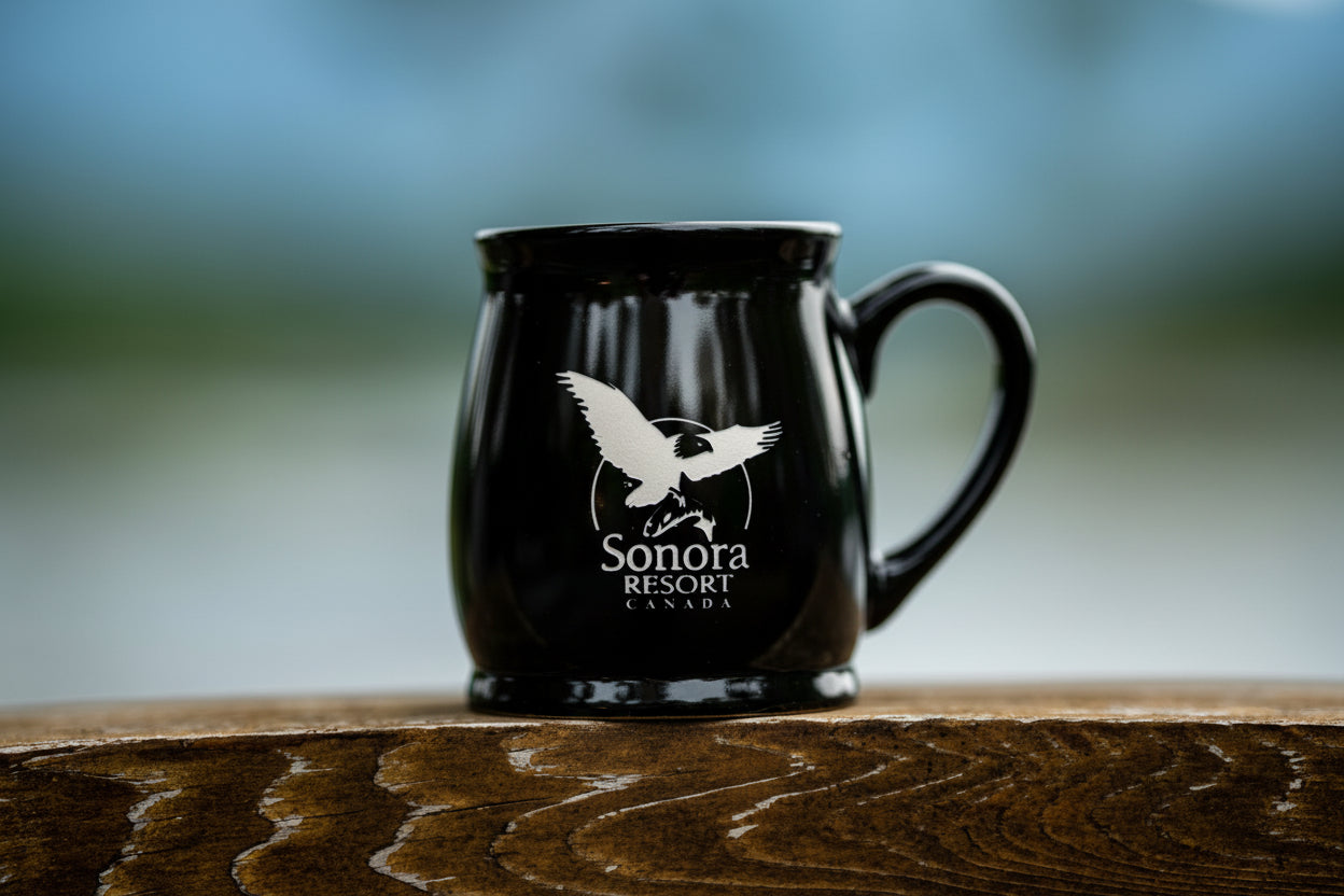 Midnight black mug with Sonora Resort Canada logo etched on the surface, sitting on a wooden beam.