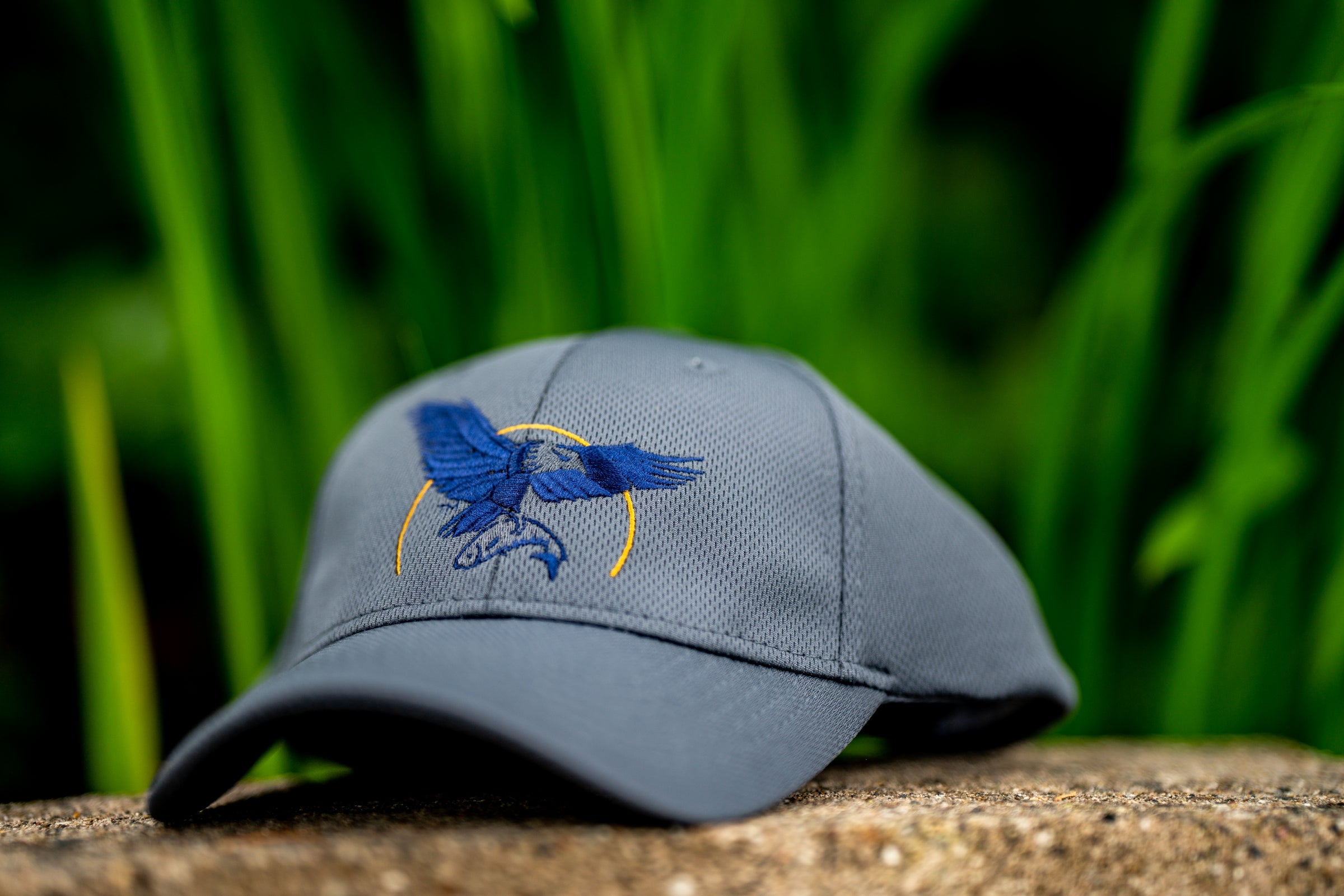 Gray ballcap with blue embroidered Sonora Resort logo on a stone surface with green grass in the background