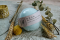 The Hobbyist botanical bath bomb in ylang ylang, lavender and cedarwood sitting on a cream blanket with grass and greenery framing it.